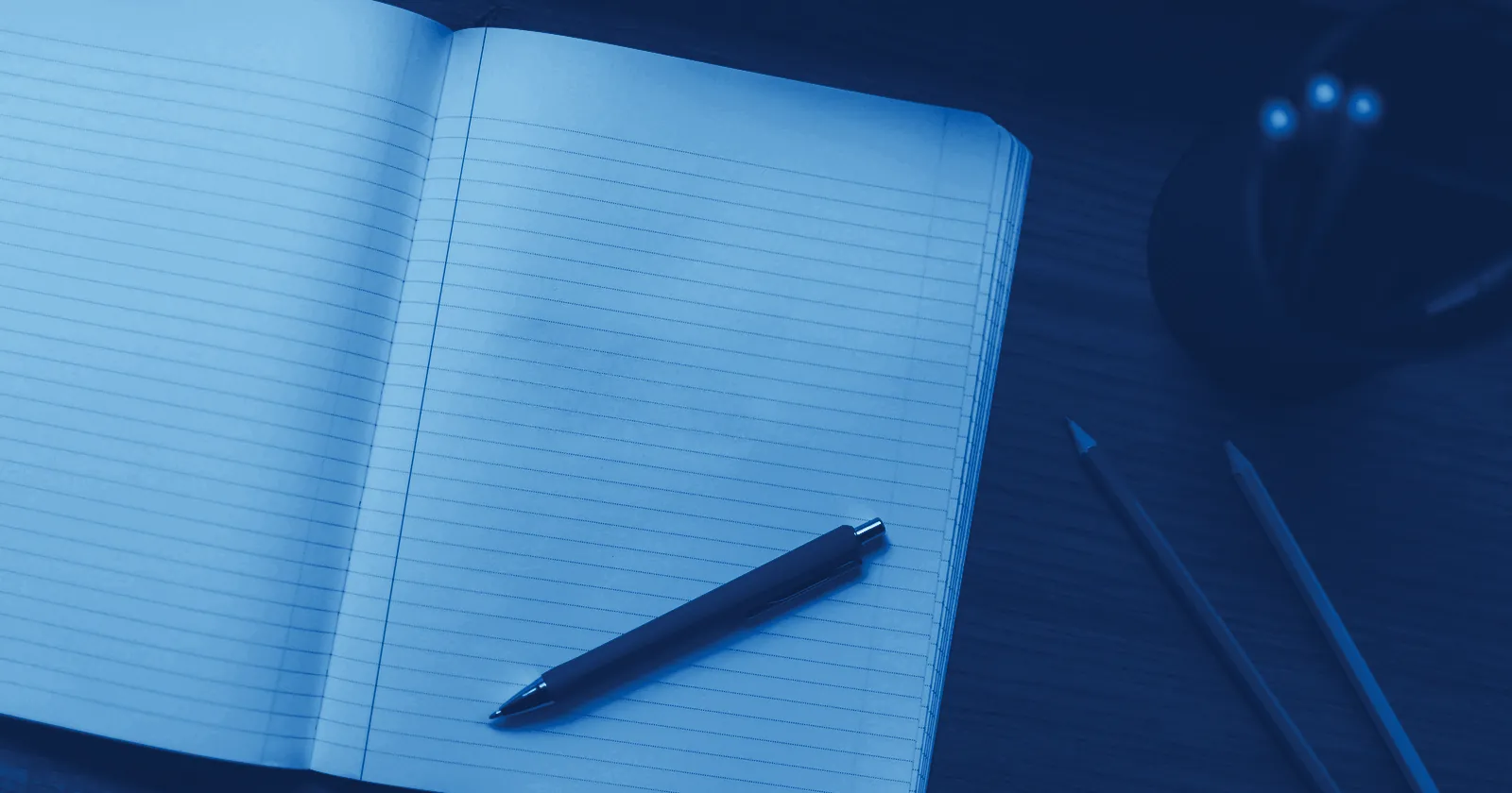 An open ruled exercise book with a pen and pencils on a desk