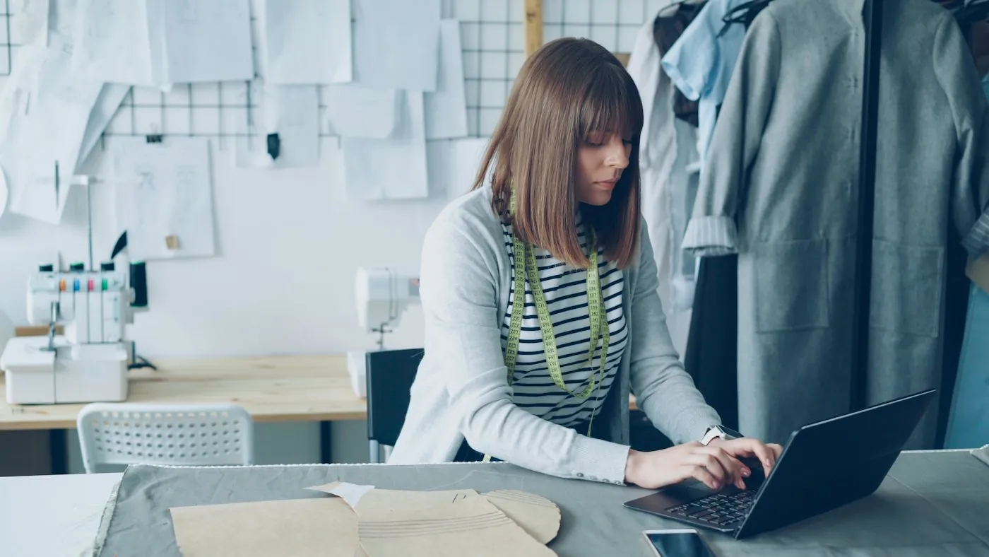 A designer at her laptop in a studio, with patterns and garments around her