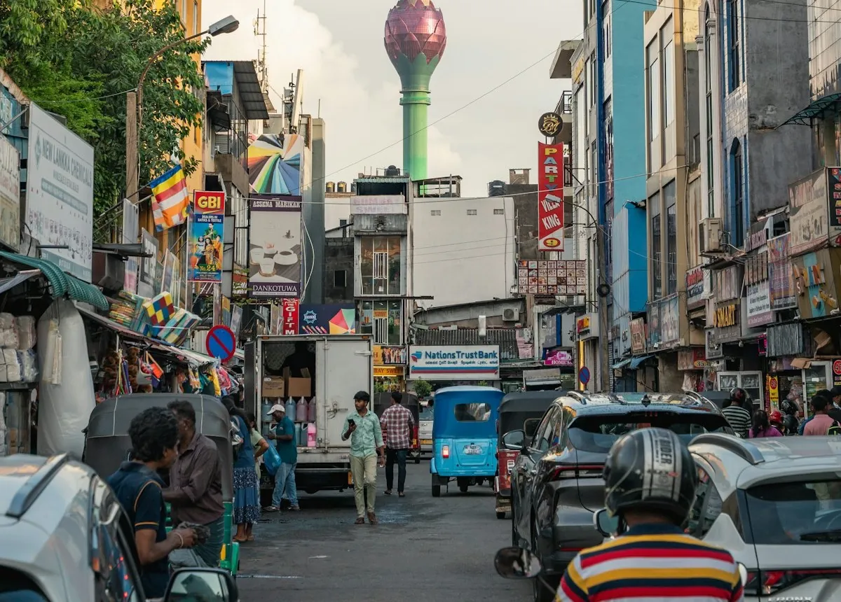 A busy street in Pettah, Colombo, with cars and shoppers and the Lotus Tower behind